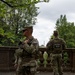 Two U.S. National Guard soldiers in full combat gear are standing on a stone terrace or balustrade, appearing to conduct a security patrol or observation post. Both wear OCP (Operational Camouflage Pattern) uniforms with American flag patches and tactical vests. In the background, a tall obelisk-shaped monument — consistent with the Washington Monument — is visible through the trees, suggesting this location is in Washington, D.C.