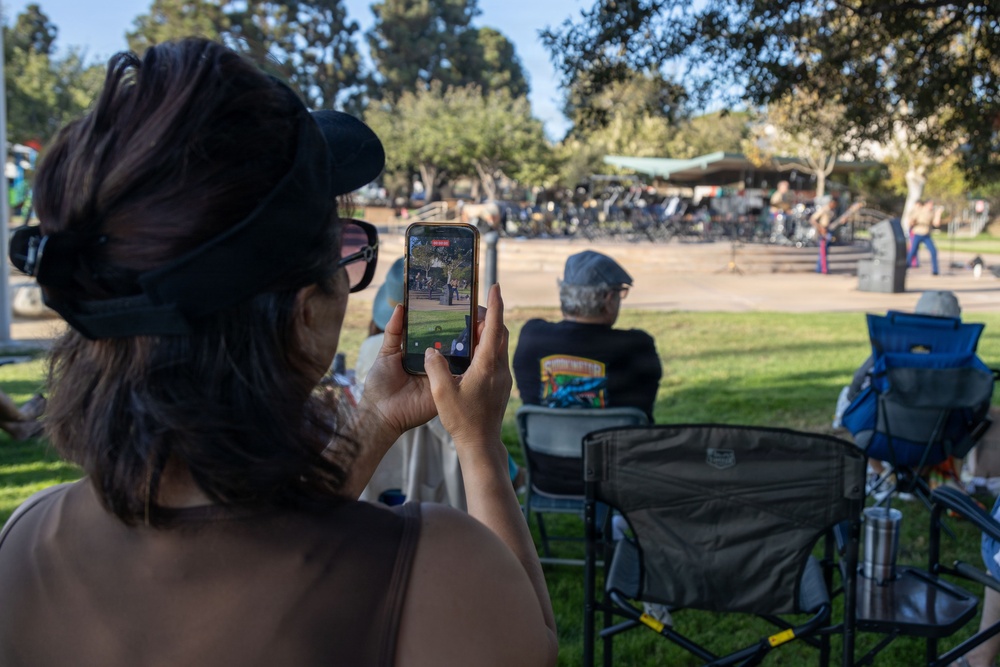 A woman wearing a black cap and sunglasses is recording a live video on her smartphone at an outdoor park event. Several people are seated in folding camp chairs on a grass lawn facing a distant stage or gathering area. The scene appears to be a casual outdoor community event on a sunny day.