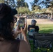 A woman wearing a black cap and sunglasses is recording a live video on her smartphone at an outdoor park event. Several people are seated in folding camp chairs on a grass lawn facing a distant stage or gathering area. The scene appears to be a casual outdoor community event on a sunny day.