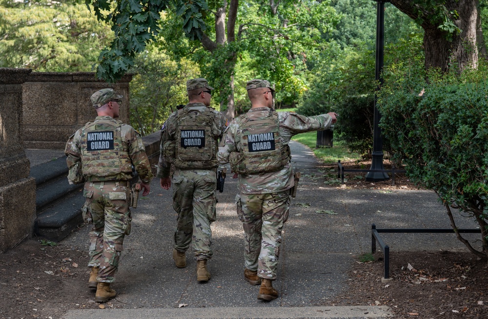 Three National Guard personnel in full tactical gear are walking along a paved pathway through a tree-lined park or garden area. All three are wearing matching multicam camouflage uniforms with 'NATIONAL GUARD' labeled tactical vests. One individual on the right appears to be pointing or gesturing in a direction, suggesting they may be conducting a patrol or reconnaissance.