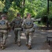 Three National Guard personnel in full tactical gear are walking along a paved pathway through a tree-lined park or garden area. All three are wearing matching multicam camouflage uniforms with 'NATIONAL GUARD' labeled tactical vests. One individual on the right appears to be pointing or gesturing in a direction, suggesting they may be conducting a patrol or reconnaissance.