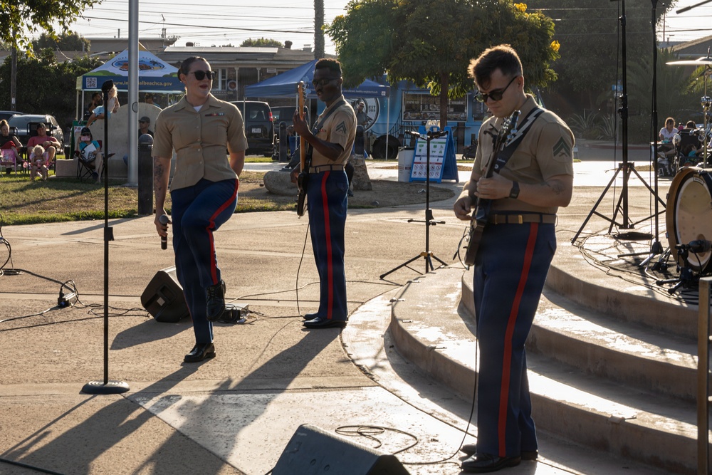 Three U.S. Marines in dress uniform are performing at an outdoor public event, likely a community concert or festival. Two are playing electric guitars while one serves as the vocalist holding a microphone. The performance takes place on a small outdoor stage or plaza with an audience visible in the background.