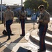 Three U.S. Marines in dress uniform are performing at an outdoor public event, likely a community concert or festival. Two are playing electric guitars while one serves as the vocalist holding a microphone. The performance takes place on a small outdoor stage or plaza with an audience visible in the background.