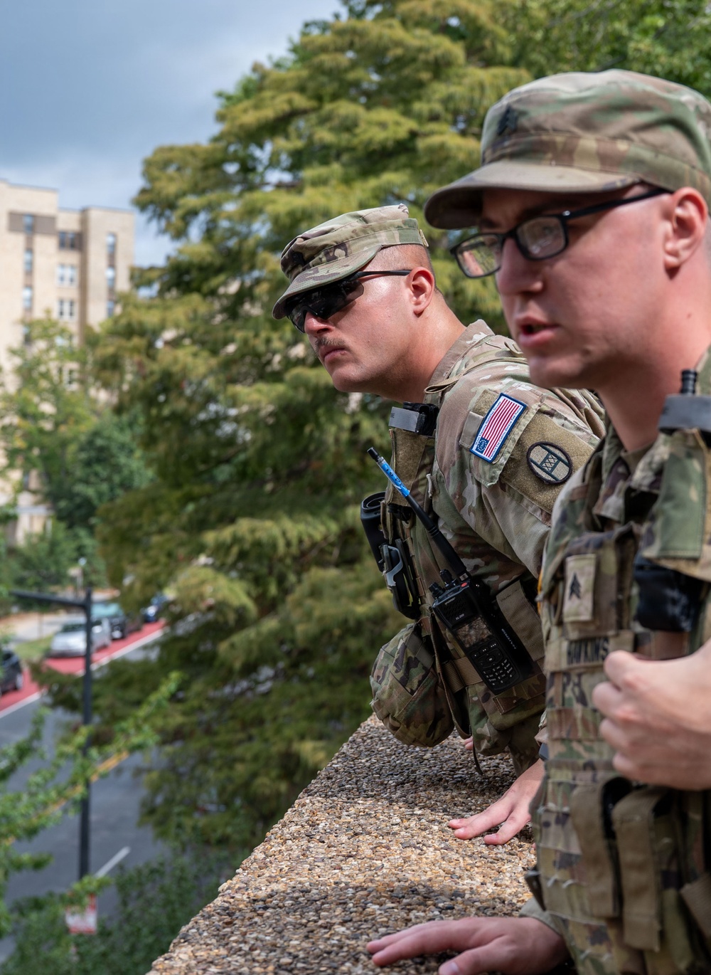 Two U.S. Army soldiers in Operational Camouflage Pattern (OCP) uniforms are positioned on an elevated rooftop or parapet, looking outward over an urban area. Both are equipped with tactical gear including radio communications devices and body armor. The setting appears to be a military installation or urban environment based on the visible buildings and street below.