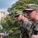 Two U.S. Army soldiers in Operational Camouflage Pattern (OCP) uniforms are positioned on an elevated rooftop or parapet, looking outward over an urban area. Both are equipped with tactical gear including radio communications devices and body armor. The setting appears to be a military installation or urban environment based on the visible buildings and street below.