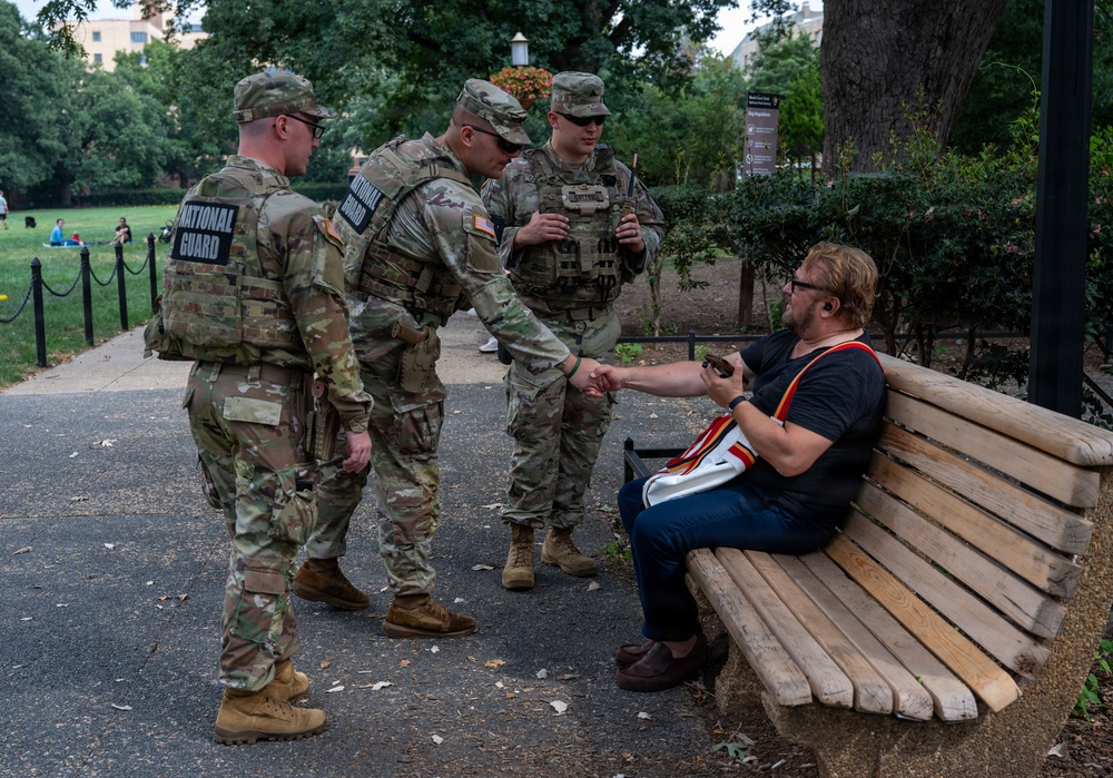 Three U.S. National Guard soldiers in camouflage uniforms and tactical vests are interacting with a civilian man seated on a wooden park bench. One soldier appears to be shaking hands or exchanging something with the seated individual. The scene takes place in an urban park setting with trees, walkways, and buildings visible in the background.
