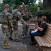 Three U.S. National Guard soldiers in camouflage uniforms and tactical vests are interacting with a civilian man seated on a wooden park bench. One soldier appears to be shaking hands or exchanging something with the seated individual. The scene takes place in an urban park setting with trees, walkways, and buildings visible in the background.
