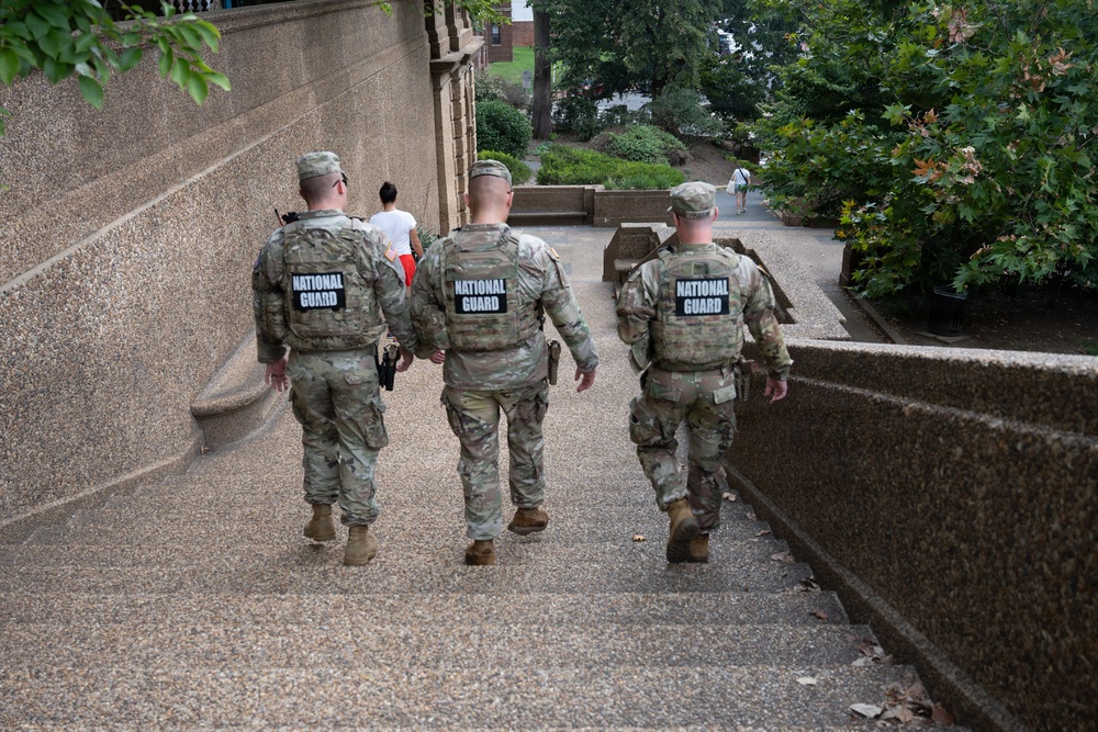 Three National Guard personnel in full military camouflage uniforms and body armor are walking away from the camera along an outdoor pathway. Each individual has a clearly visible 'NATIONAL GUARD' patch on the back of their tactical vest. The setting appears to be a public area with stone architecture, trees, and a few civilians visible in the background.
