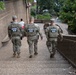 Three National Guard personnel in full military camouflage uniforms and body armor are walking away from the camera along an outdoor pathway. Each individual has a clearly visible 'NATIONAL GUARD' patch on the back of their tactical vest. The setting appears to be a public area with stone architecture, trees, and a few civilians visible in the background.