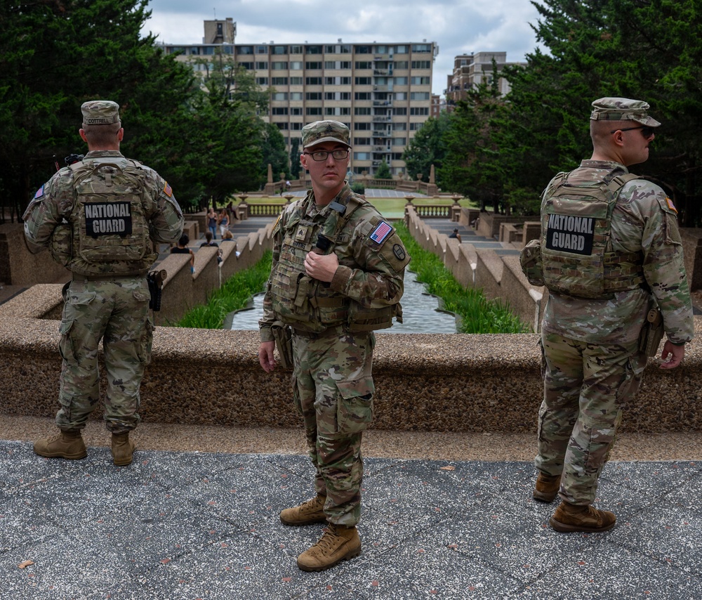 Three U.S. National Guard soldiers in full combat gear are standing at what appears to be a public outdoor space, likely in Washington D.C., based on the architectural features visible in the background. Two soldiers have their backs to the camera with 'NATIONAL GUARD' clearly visible on their tactical vests, while the center soldier faces forward wearing glasses and holding a radio. The setting includes a decorative water feature or reflecting pool with terraced stonework and urban buildings in the background.