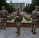 Three U.S. National Guard soldiers in full combat gear are standing at what appears to be a public outdoor space, likely in Washington D.C., based on the architectural features visible in the background. Two soldiers have their backs to the camera with 'NATIONAL GUARD' clearly visible on their tactical vests, while the center soldier faces forward wearing glasses and holding a radio. The setting includes a decorative water feature or reflecting pool with terraced stonework and urban buildings in the background.