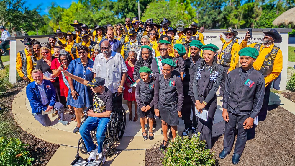 A large group photo taken outdoors at what appears to be a military memorial or monument dedication ceremony. The group includes adults and youth wearing distinctive uniforms — many in yellow vests with black cowboy hats, and younger participants in black attire with green berets. A person in a wheelchair is prominently positioned in the foreground.