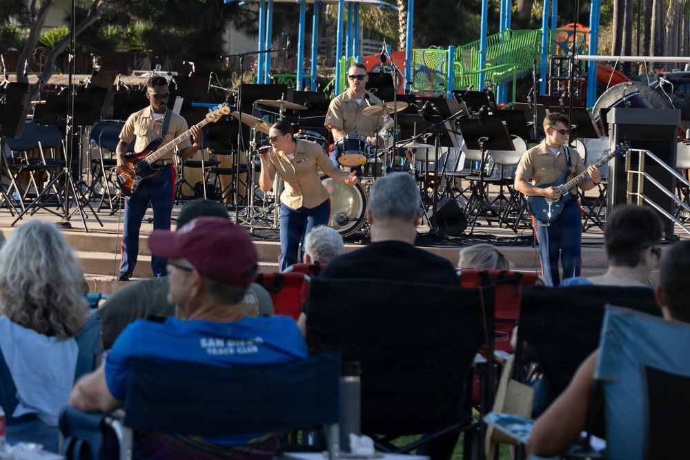 A military band, likely U.S. Marines based on their tan and blue uniforms, performs an outdoor concert on a stage in what appears to be a public park. Four visible performers include a bassist, a vocalist, a drummer, and a guitarist, all wearing matching military dress uniforms. An audience of civilians is seated in folding chairs watching the performance in what appears to be late afternoon sunlight.