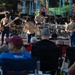 A military band, likely U.S. Marines based on their tan and blue uniforms, performs an outdoor concert on a stage in what appears to be a public park. Four visible performers include a bassist, a vocalist, a drummer, and a guitarist, all wearing matching military dress uniforms. An audience of civilians is seated in folding chairs watching the performance in what appears to be late afternoon sunlight.