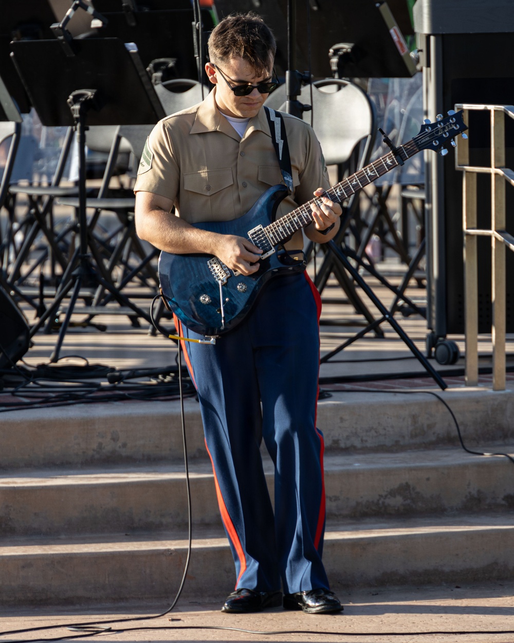 A young male in U.S. Marine Corps uniform is playing an electric guitar on an outdoor stage or plaza. He wears the distinctive khaki shirt and blue trousers with red 'blood stripe' of Marine dress uniform, paired with black sunglasses. Multiple music stands and equipment are visible in the background, indicating a band or ensemble performance setting.