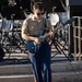 A young male in U.S. Marine Corps uniform is playing an electric guitar on an outdoor stage or plaza. He wears the distinctive khaki shirt and blue trousers with red 'blood stripe' of Marine dress uniform, paired with black sunglasses. Multiple music stands and equipment are visible in the background, indicating a band or ensemble performance setting.