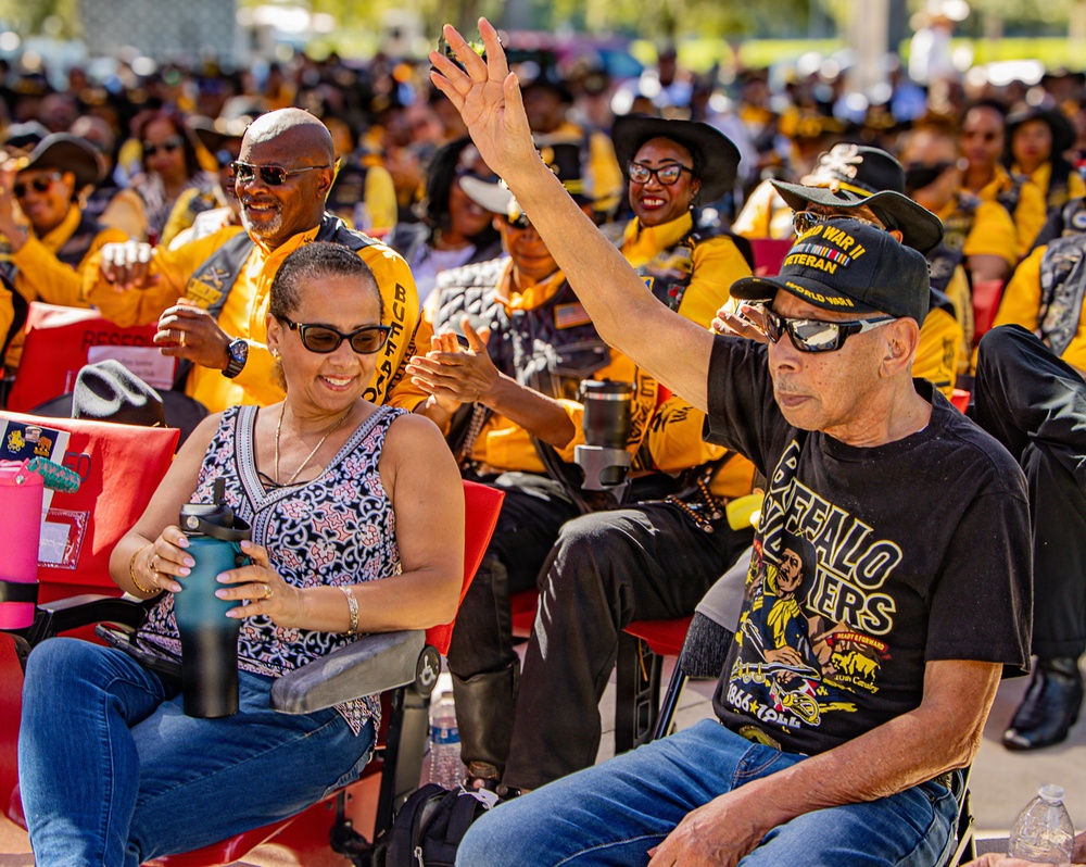 A large outdoor gathering featuring a crowd predominantly wearing black and gold/yellow uniforms or jackets, likely a Buffalo Soldiers organization event or reunion. In the foreground, a man wearing a 'Buffalo Soldiers' t-shirt and a 'World War II Veteran' cap raises his hand, seated next to a woman holding a water bottle. The atmosphere appears celebratory and engaged, with attendees clapping and smiling.