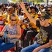 A large outdoor gathering featuring a crowd predominantly wearing black and gold/yellow uniforms or jackets, likely a Buffalo Soldiers organization event or reunion. In the foreground, a man wearing a 'Buffalo Soldiers' t-shirt and a 'World War II Veteran' cap raises his hand, seated next to a woman holding a water bottle. The atmosphere appears celebratory and engaged, with attendees clapping and smiling.