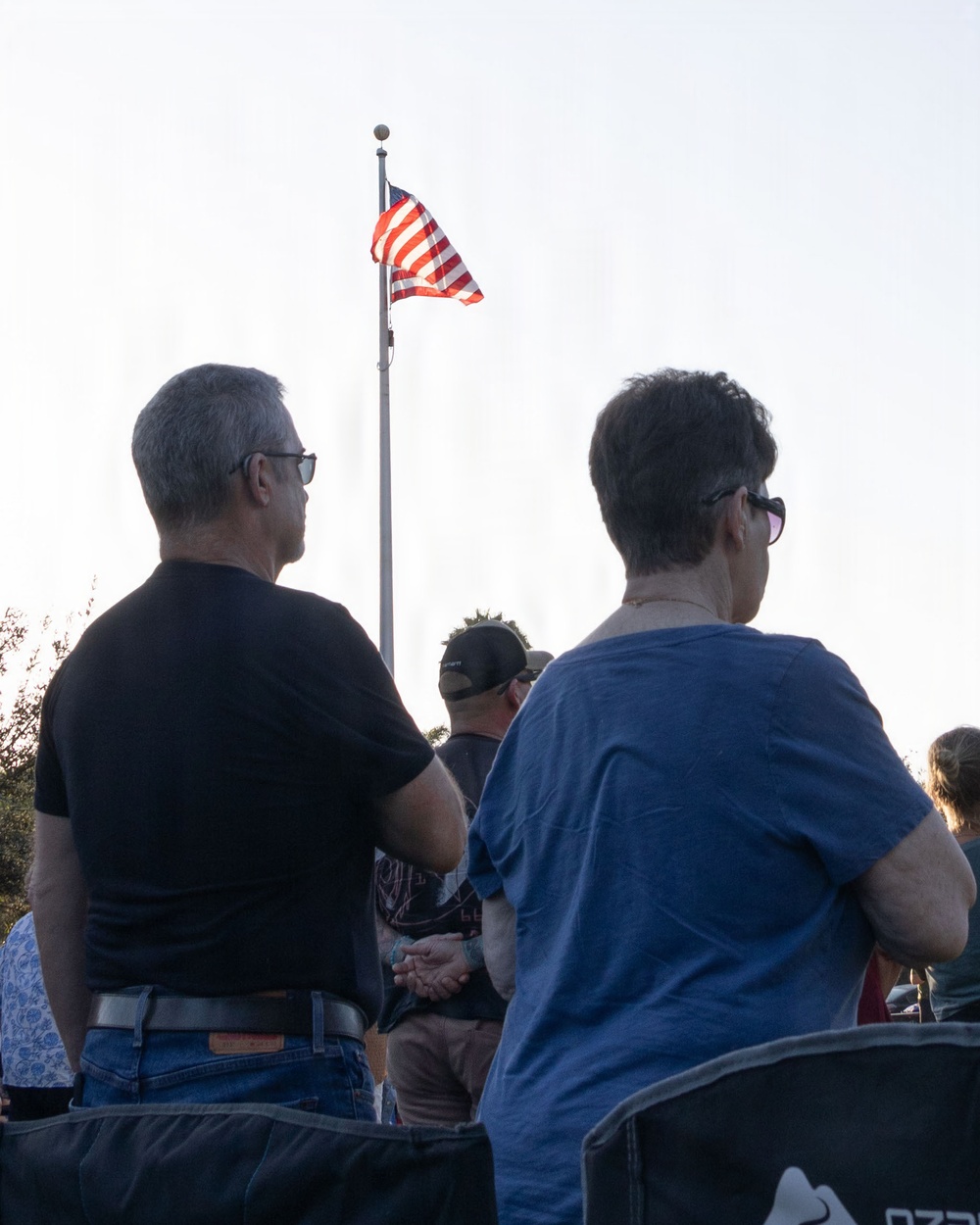 A group of people are gathered outdoors facing an American flag on a tall flagpole. The scene appears to be a patriotic or commemorative event, possibly a ceremony, with attendees standing and facing the flag. The photo is taken from behind the crowd during what appears to be late afternoon or early evening light.