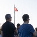A group of people are gathered outdoors facing an American flag on a tall flagpole. The scene appears to be a patriotic or commemorative event, possibly a ceremony, with attendees standing and facing the flag. The photo is taken from behind the crowd during what appears to be late afternoon or early evening light.