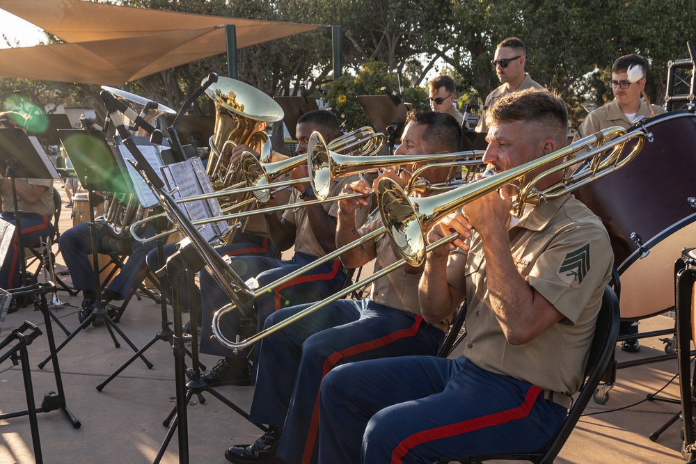 A military band, identifiable as U.S. Marines by their distinctive tan shirts and blue trousers with red stripes, performs an outdoor concert. Multiple musicians are playing trombones in the foreground, with additional brass and percussion instruments visible in the background. The performance appears to take place in a shaded outdoor venue during golden hour lighting.