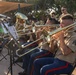 A military band, identifiable as U.S. Marines by their distinctive tan shirts and blue trousers with red stripes, performs an outdoor concert. Multiple musicians are playing trombones in the foreground, with additional brass and percussion instruments visible in the background. The performance appears to take place in a shaded outdoor venue during golden hour lighting.