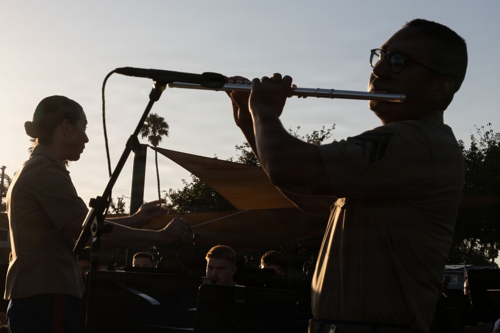 An outdoor musical performance captured at dusk or golden hour, showing silhouetted figures against a bright sky. A person on the right is playing a flute while wearing glasses, and another person on the left appears to be playing a guitar or similar stringed instrument. Several audience members are visible seated in the background.