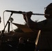 An outdoor musical performance captured at dusk or golden hour, showing silhouetted figures against a bright sky. A person on the right is playing a flute while wearing glasses, and another person on the left appears to be playing a guitar or similar stringed instrument. Several audience members are visible seated in the background.