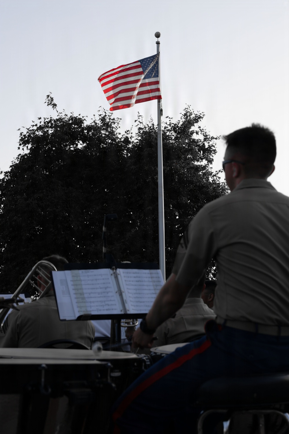 An outdoor military or patriotic band performance is taking place with musicians in tan/khaki uniforms seated with sheet music on stands. An American flag flies prominently on a flagpole in the background against a light sky, with a large tree silhouetted behind it. The scene appears to be a formal outdoor concert or ceremony setting.