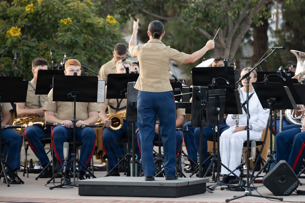 A military band performance is taking place outdoors, conducted by a female officer standing on a podium with her back to the camera. The musicians, dressed in U.S. Marine Corps and U.S. Navy uniforms, are seated with music stands and playing various instruments. The setting appears to be a public outdoor venue with trees visible in the background.