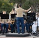 A military band performance is taking place outdoors, conducted by a female officer standing on a podium with her back to the camera. The musicians, dressed in U.S. Marine Corps and U.S. Navy uniforms, are seated with music stands and playing various instruments. The setting appears to be a public outdoor venue with trees visible in the background.