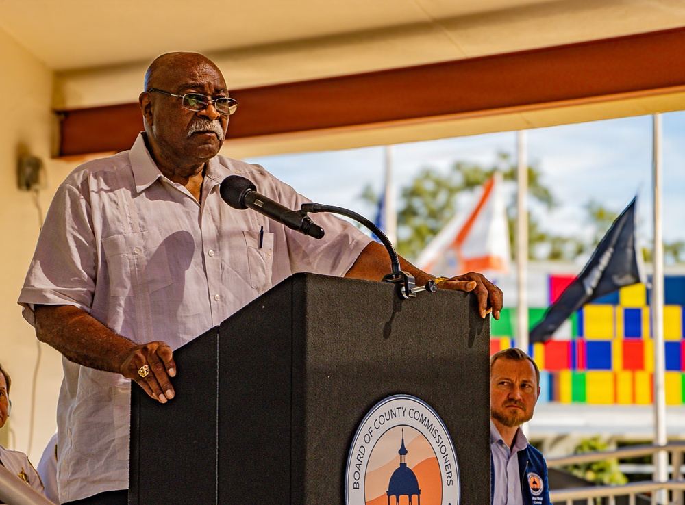 An older man is speaking at a podium labeled 'Board of County Commissioners' at what appears to be an outdoor public event. He is wearing a light-colored button-up shirt and glasses, and is positioned in front of a microphone. In the background, colorful flags, a dark flag, and another attendee are visible.