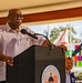 An older man is speaking at a podium labeled 'Board of County Commissioners' at what appears to be an outdoor public event. He is wearing a light-colored button-up shirt and glasses, and is positioned in front of a microphone. In the background, colorful flags, a dark flag, and another attendee are visible.