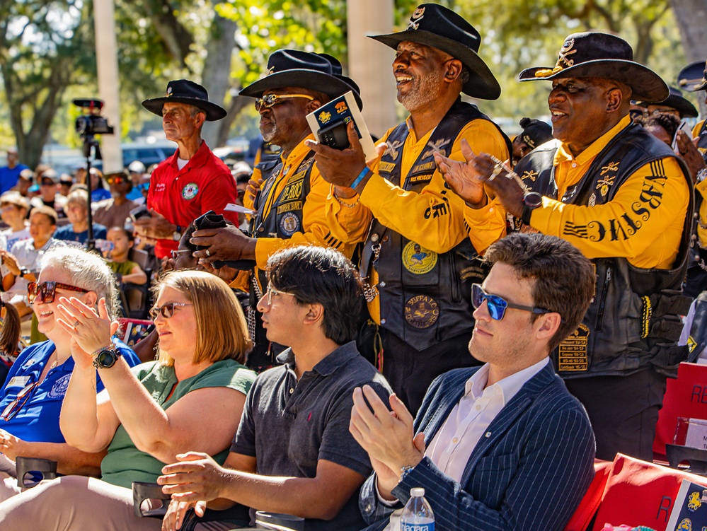 An outdoor public event or ceremony featuring a group of men wearing matching black and gold vests with patches and wide-brimmed black cavalry-style hats, consistent with Buffalo Soldiers motorcycle club or commemorative organization attire. They are standing and applauding enthusiastically among a seated crowd of diverse attendees. The setting appears to be a daytime outdoor gathering under trees with a large audience in the background.