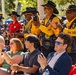 An outdoor public event or ceremony featuring a group of men wearing matching black and gold vests with patches and wide-brimmed black cavalry-style hats, consistent with Buffalo Soldiers motorcycle club or commemorative organization attire. They are standing and applauding enthusiastically among a seated crowd of diverse attendees. The setting appears to be a daytime outdoor gathering under trees with a large audience in the background.