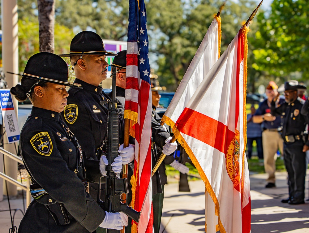 A Hillsborough County Sheriff's Office color guard is presenting flags at an outdoor ceremony. Officers in formal dress uniforms carry the American flag and the Florida state flag, with rifles at ceremonial positions. Additional law enforcement personnel and civilians are visible in the background.