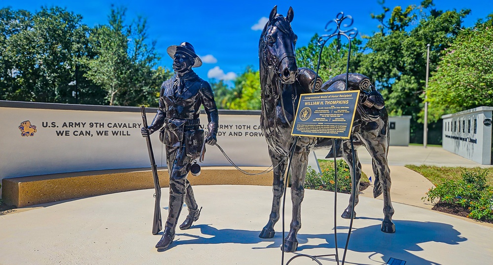 A bronze statue memorial depicting a Buffalo Soldier leading a horse, dedicated to Congressional Medal of Honor Recipient William H. Thompkins. The monument is part of a larger memorial complex honoring the U.S. Army 9th and 10th Cavalry regiments, known as the Buffalo Soldiers. The setting features a curved white wall with unit mottos and insignia, surrounded by lush green trees under a bright blue sky.