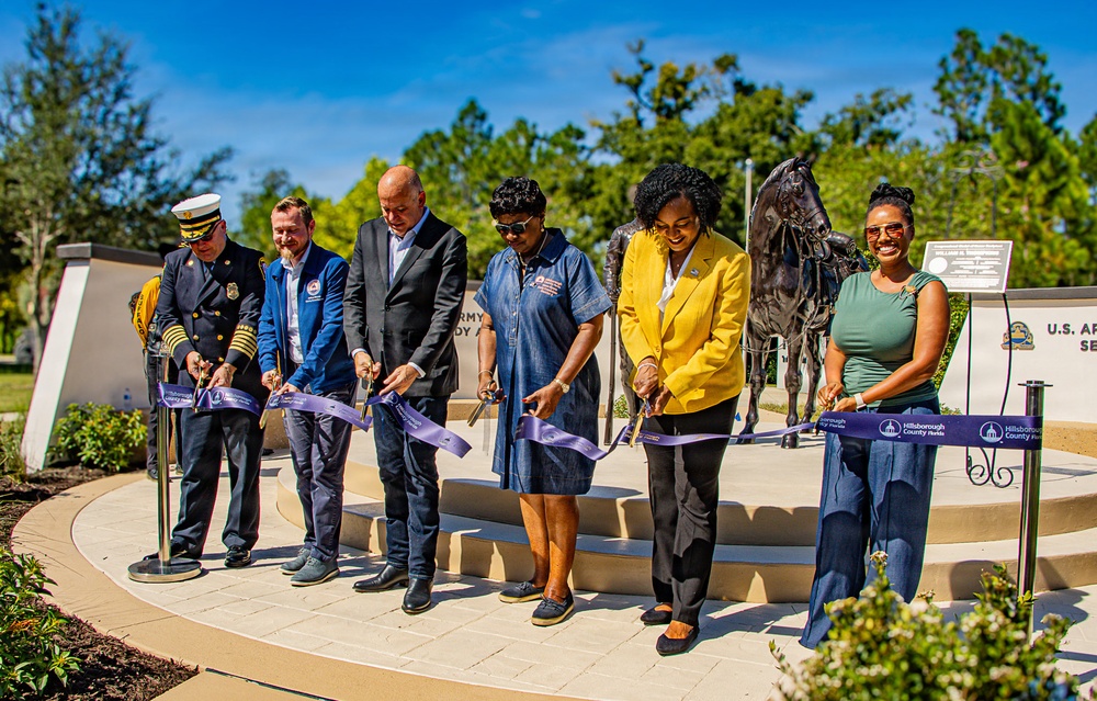 A ribbon-cutting ceremony is taking place at what appears to be a U.S. Army memorial or service location in Hillsborough County, Florida. Six individuals, including a uniformed fire official and several dignitaries, are cutting a purple ribbon simultaneously. A bronze equestrian statue is visible in the background, suggesting this is a dedication or grand opening event.