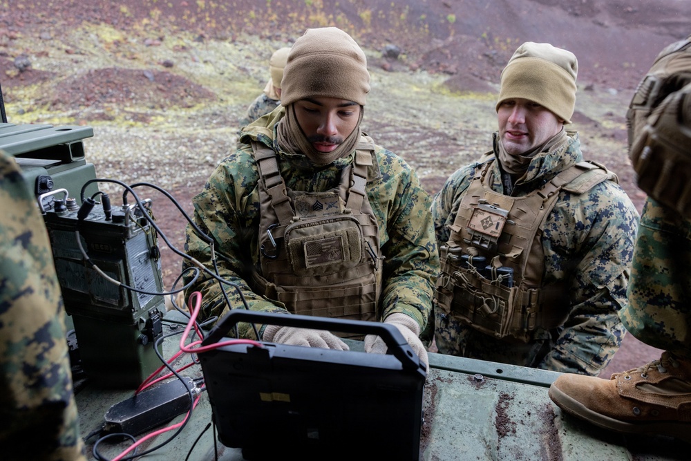 Several military personnel in MARPAT (Marine Pattern) camouflage uniforms and tactical gear are gathered around a ruggedized laptop computer connected to military communications equipment. They appear to be conducting field operations or training in a rugged, volcanic-terrain environment. The soldiers are wearing cold-weather gear including tan fleece caps and gloves, suggesting operations in a cold climate.
