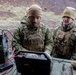 Several military personnel in MARPAT (Marine Pattern) camouflage uniforms and tactical gear are gathered around a ruggedized laptop computer connected to military communications equipment. They appear to be conducting field operations or training in a rugged, volcanic-terrain environment. The soldiers are wearing cold-weather gear including tan fleece caps and gloves, suggesting operations in a cold climate.