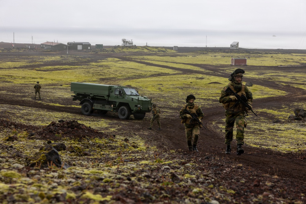 Military personnel in camouflage uniforms are conducting a patrol or exercise across a volcanic, moss-covered landscape, accompanied by a green military truck. The terrain appears to be characteristic of Iceland or a similar North Atlantic volcanic region, with dark basaltic soil and bright yellow-green moss. Several soldiers are moving on foot along a dirt track while a military vehicle follows behind.
