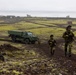Military personnel in camouflage uniforms are conducting a patrol or exercise across a volcanic, moss-covered landscape, accompanied by a green military truck. The terrain appears to be characteristic of Iceland or a similar North Atlantic volcanic region, with dark basaltic soil and bright yellow-green moss. Several soldiers are moving on foot along a dirt track while a military vehicle follows behind.