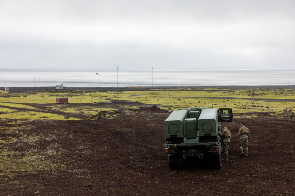 Two military personnel in camouflage uniforms are standing beside a large green military vehicle, identified as an M142 HIMARS (High Mobility Artillery Rocket System) or similar rocket artillery launcher, positioned on dark volcanic terrain. The landscape features moss-covered lava fields with a coastal shoreline visible in the background under an overcast sky. Small structures, including a red building, are visible in the mid-distance.