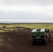 Two military personnel in camouflage uniforms are standing beside a large green military vehicle, identified as an M142 HIMARS (High Mobility Artillery Rocket System) or similar rocket artillery launcher, positioned on dark volcanic terrain. The landscape features moss-covered lava fields with a coastal shoreline visible in the background under an overcast sky. Small structures, including a red building, are visible in the mid-distance.