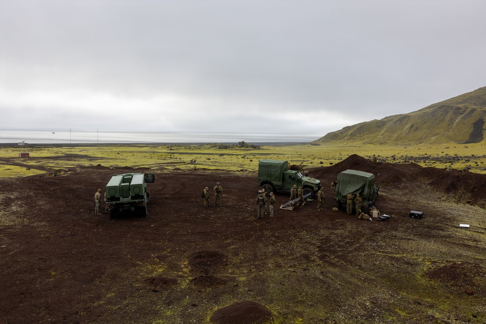 A group of military personnel are gathered around three green military vehicles on a dark volcanic terrain, likely during a field exercise or operational deployment. The landscape features reddish-brown volcanic soil with patches of green moss or lichen, a flat coastal plain in the background, and a green hillside to the right. The overcast sky and coastal water visible in the distance suggest a northern or subarctic environment.