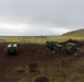 A group of military personnel are gathered around three green military vehicles on a dark volcanic terrain, likely during a field exercise or operational deployment. The landscape features reddish-brown volcanic soil with patches of green moss or lichen, a flat coastal plain in the background, and a green hillside to the right. The overcast sky and coastal water visible in the distance suggest a northern or subarctic environment.