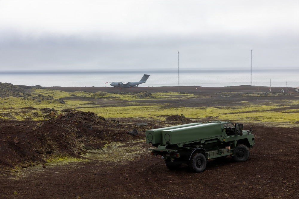 A military rocket artillery launcher vehicle, consistent with an M270 MLRS or similar system, is positioned on dark volcanic terrain with green moss coverage. In the background, a large military transport aircraft, consistent with a C-17 Globemaster III, is visible on or near a runway adjacent to a body of water. The setting appears to be a remote coastal military installation, likely in Iceland or a similar North Atlantic volcanic landscape.