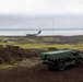 A military rocket artillery launcher vehicle, consistent with an M270 MLRS or similar system, is positioned on dark volcanic terrain with green moss coverage. In the background, a large military transport aircraft, consistent with a C-17 Globemaster III, is visible on or near a runway adjacent to a body of water. The setting appears to be a remote coastal military installation, likely in Iceland or a similar North Atlantic volcanic landscape.