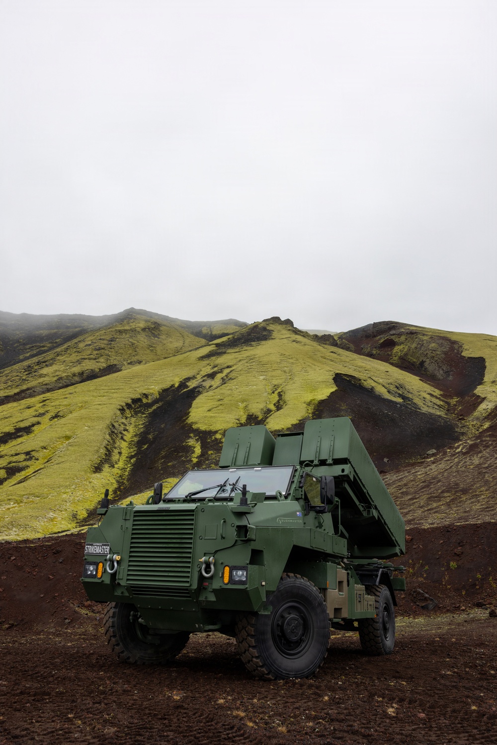 A military wheeled vehicle designated 'STRIKEMASTER' is parked on dark volcanic terrain against a backdrop of moss-covered green hills under an overcast sky. The vehicle appears to be a modern multiple launch rocket system (MLRS) or artillery platform mounted on an armored wheeled chassis, painted in olive/military green. The landscape strongly resembles Iceland's volcanic highland terrain.