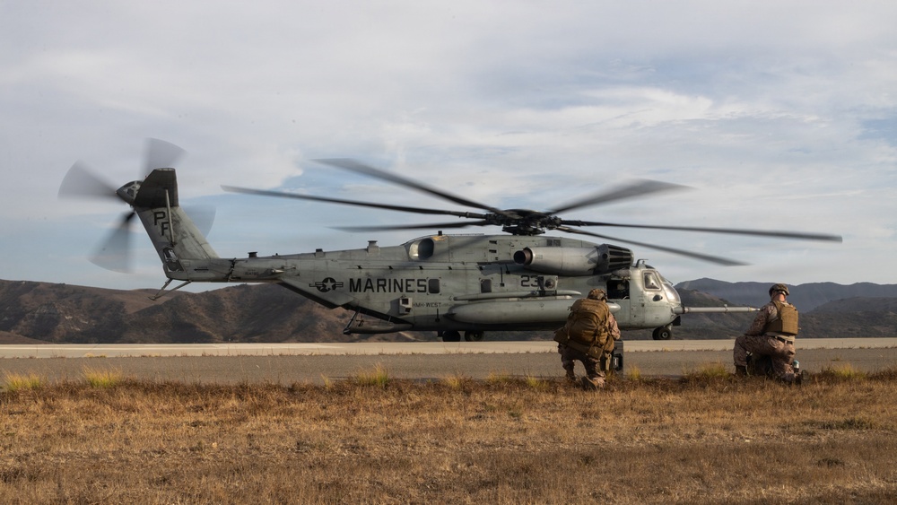 A U.S. Marines heavy-lift helicopter, identified as a Sikorsky CH-53E Super Stallion marked 'MARINES' with tail number 23, is on or near a tarmac with rotors spinning. Two Marines in combat gear are crouching nearby, appearing to wait for or prepare to board the aircraft. The setting appears to be a military training area with arid hills in the background.